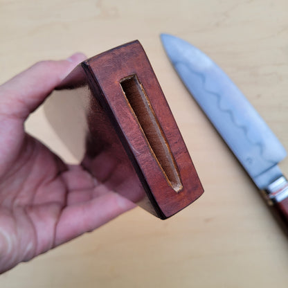 A hand holding a dark reddish-brown wooden knife block with a recessed slot, with a partially visible chef's knife in the background on a light wood surface.