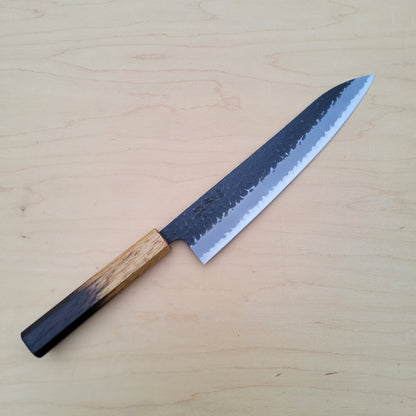 A Japanese-style kitchen knife with a dark textured steel blade and a light brown wooden handle, placed diagonally on a light beige wood grain surface.
