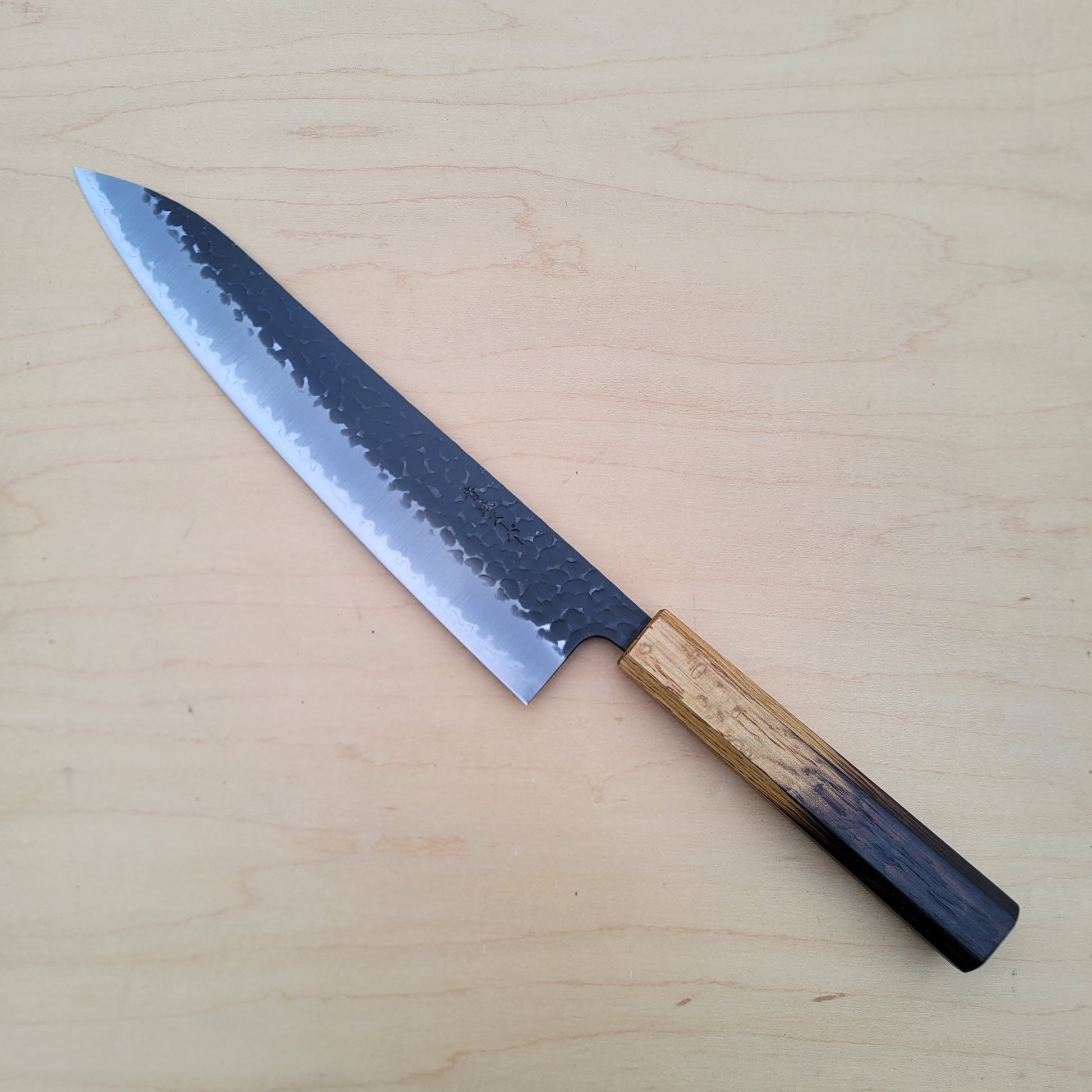 A Japanese-style kitchen knife with a hammered steel blade and a natural wood handle, placed diagonally on a light wood grain surface.