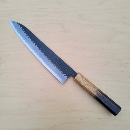 A Japanese-style kitchen knife with a hammered steel blade and a natural wood handle, placed diagonally on a light wood grain surface.