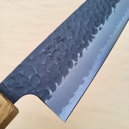 A close-up of a Japanese-style kitchen knife with a hammered steel blade and a light wood handle, set against a light wood background.