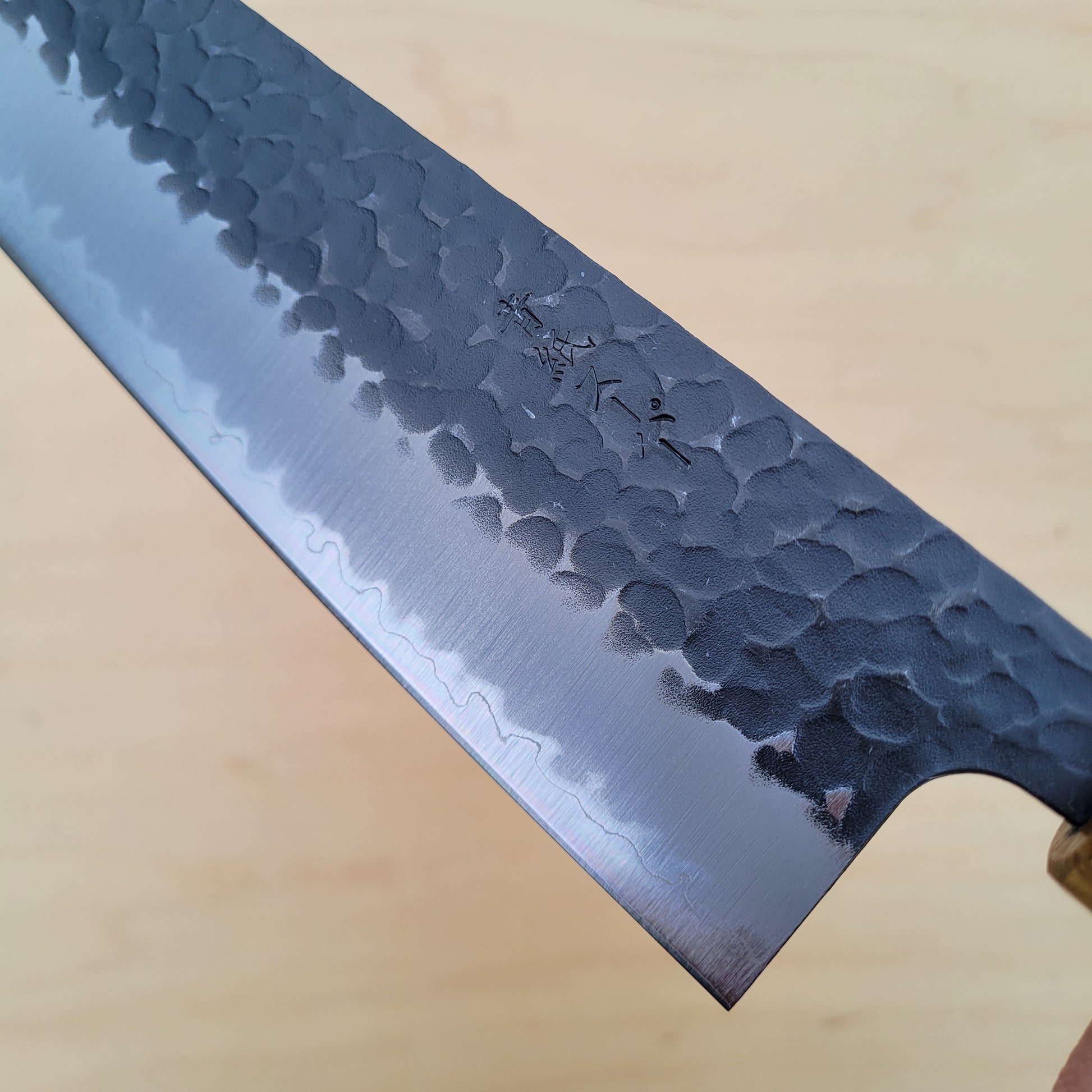 A close-up of a Japanese-style kitchen knife with a hammered and satin-finished steel blade, resting diagonally on a light wood surface.