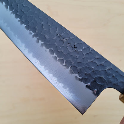 A close-up of a Japanese-style kitchen knife with a hammered and satin-finished steel blade, resting diagonally on a light wood surface.